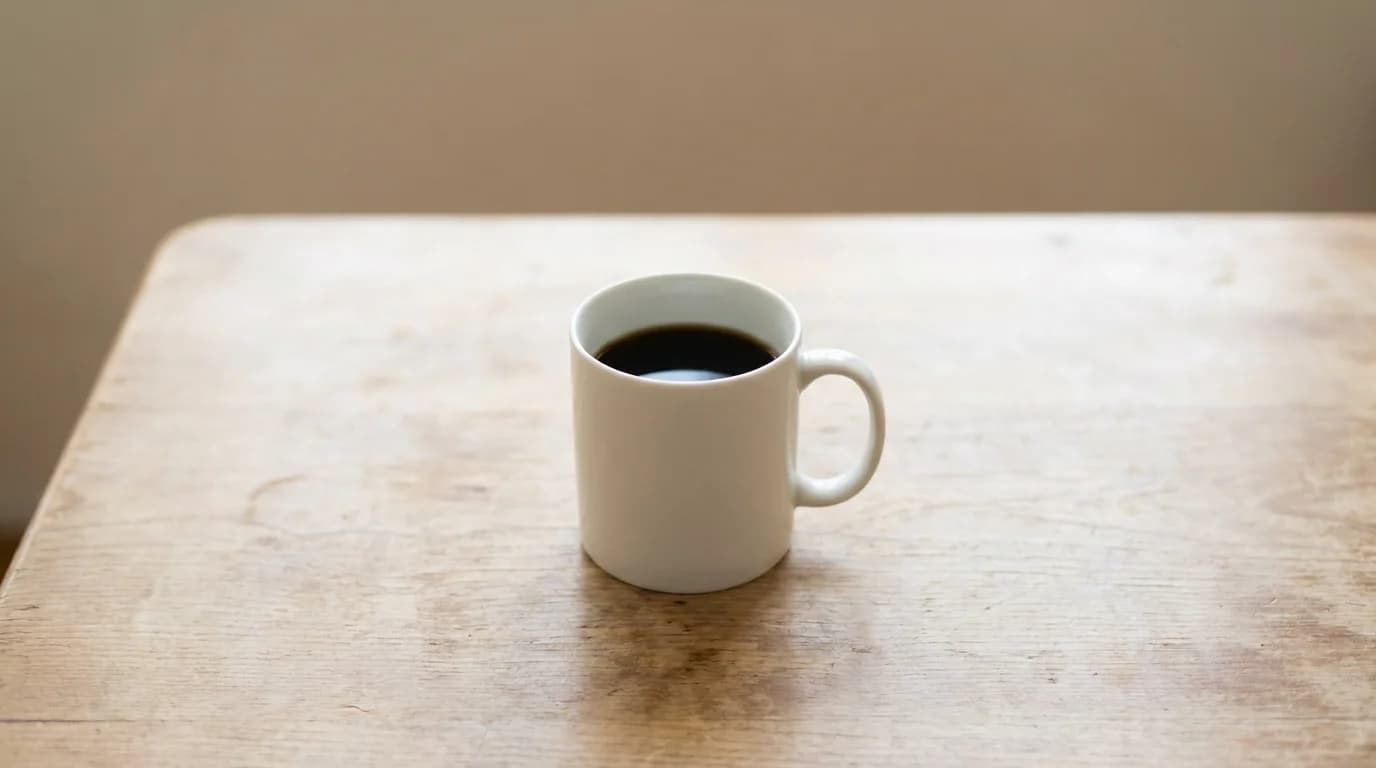 A single white ceramic mug of coffee on a bare wooden surface with soft daylight, minimalist composition