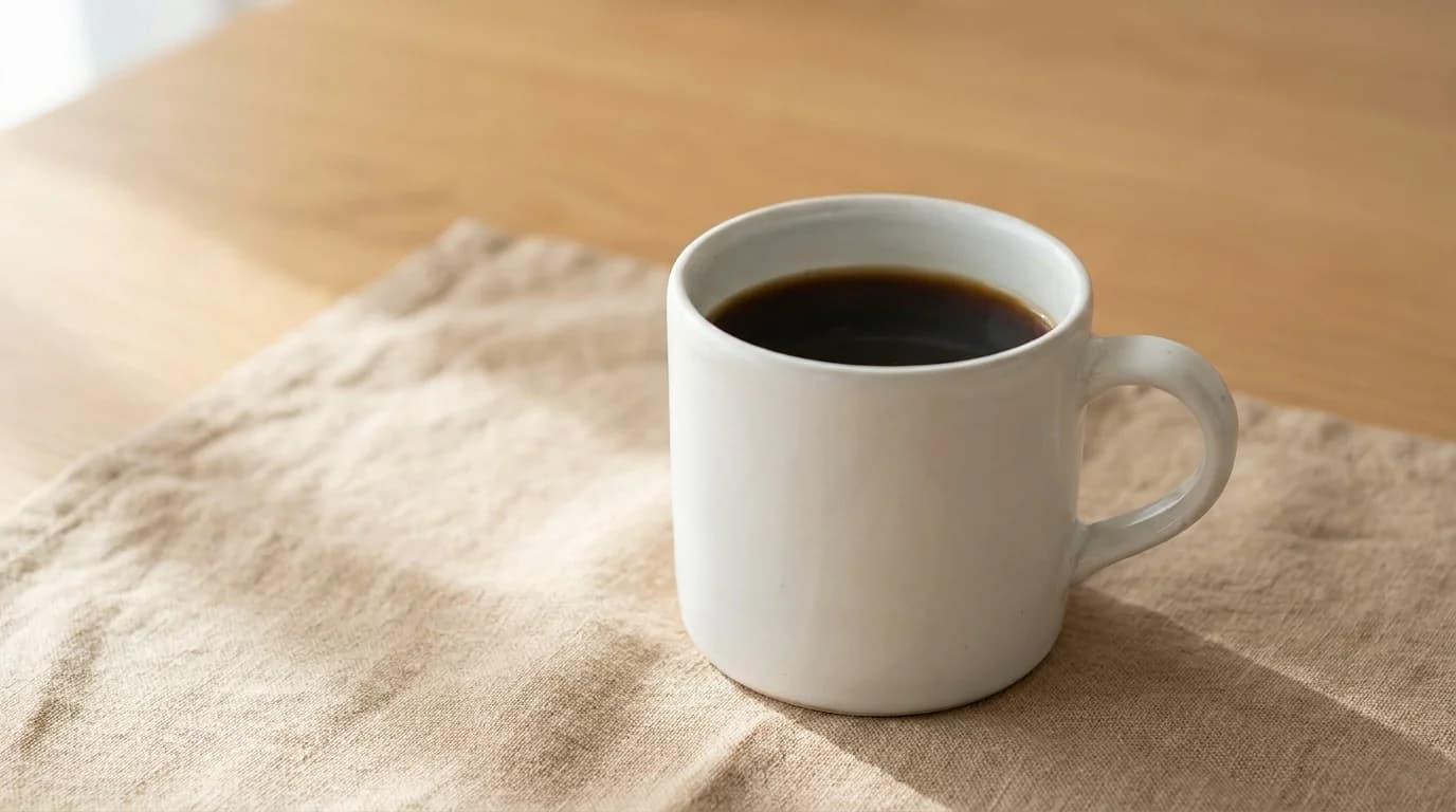 A white ceramic mug of black coffee on a bare linen surface, soft morning window light, calm minimalist composition