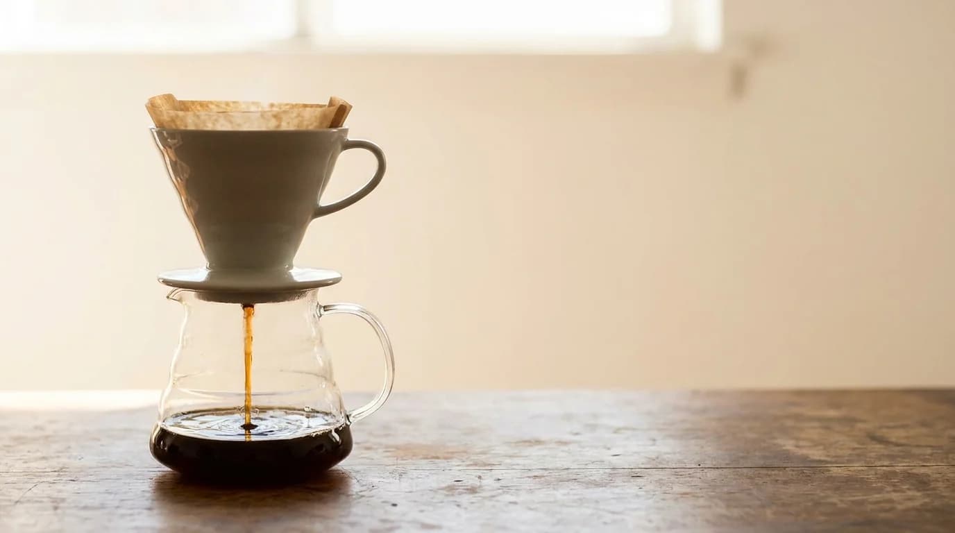 A pour-over coffee dripper on a clear glass carafe, coffee slowly extracting through a paper filter, soft daylight from behind creating a warm glow, minimalist composition