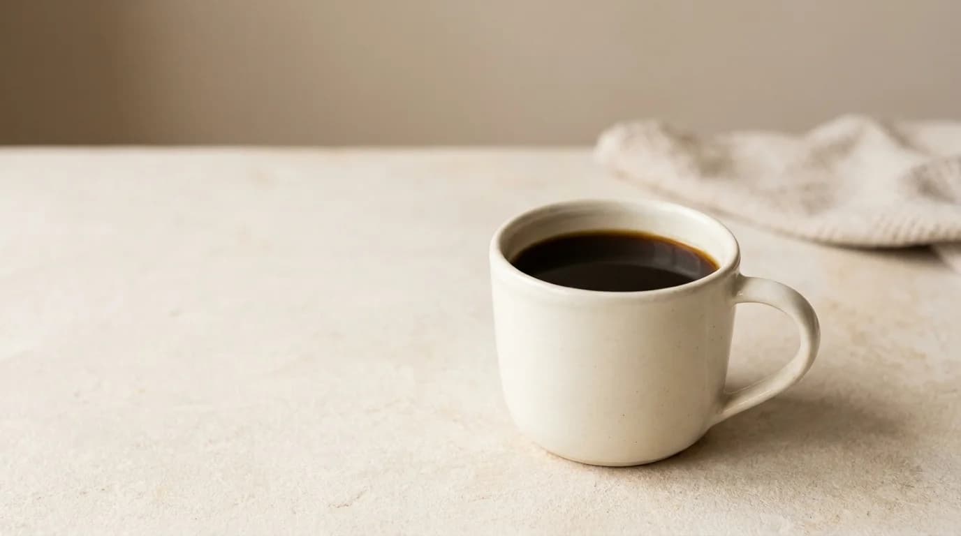 A single white ceramic mug of black coffee on a bare pale stone surface, soft diffused daylight from above, minimalist composition