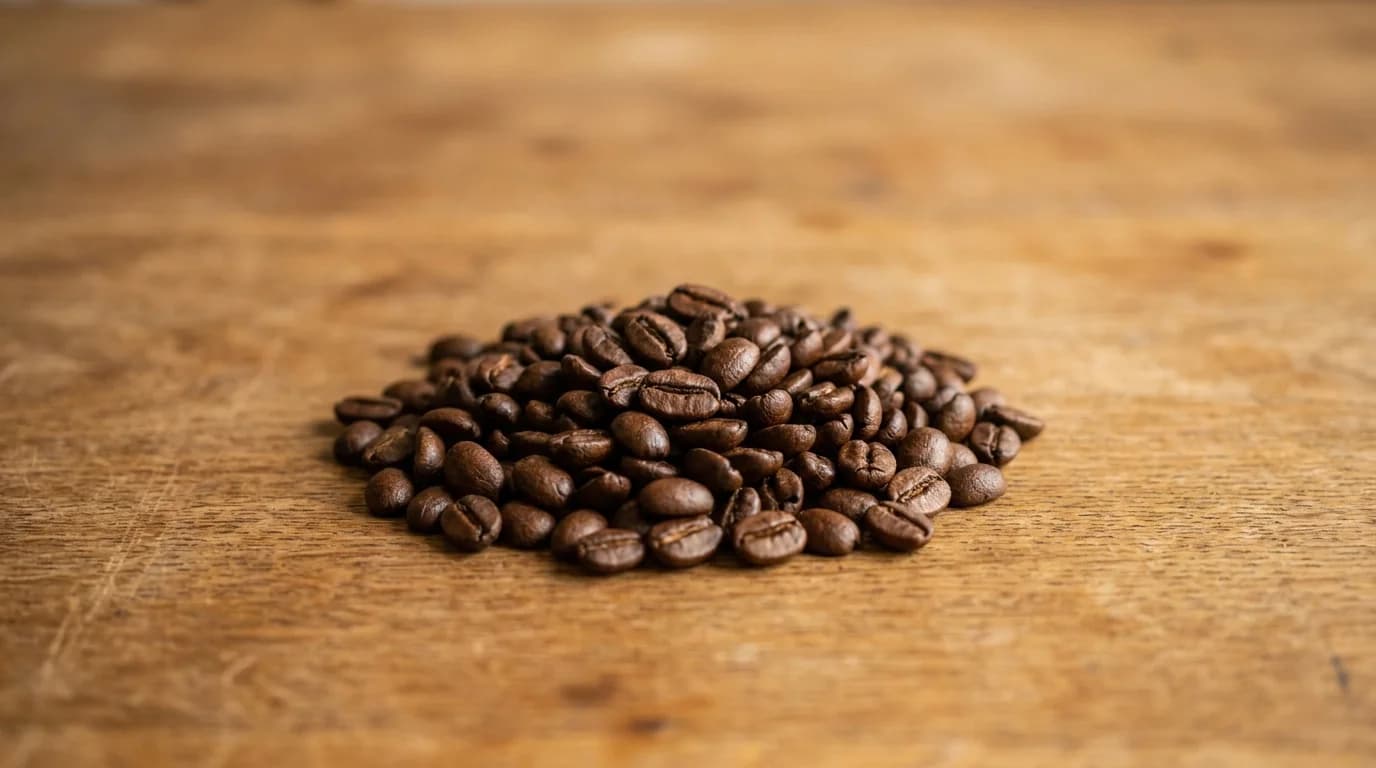 A small pile of dark medium-roasted brown coffee beans on a bare wooden surface with warm daylight from above, minimalist composition