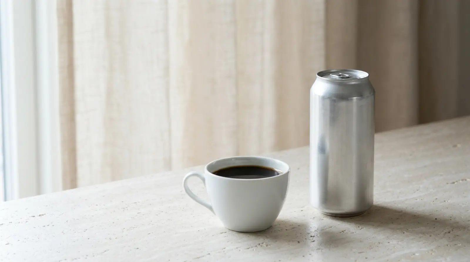 A plain white cup of black coffee and a tall unbranded aluminium can side by side on a pale counter in even daylight, no labels visible on either