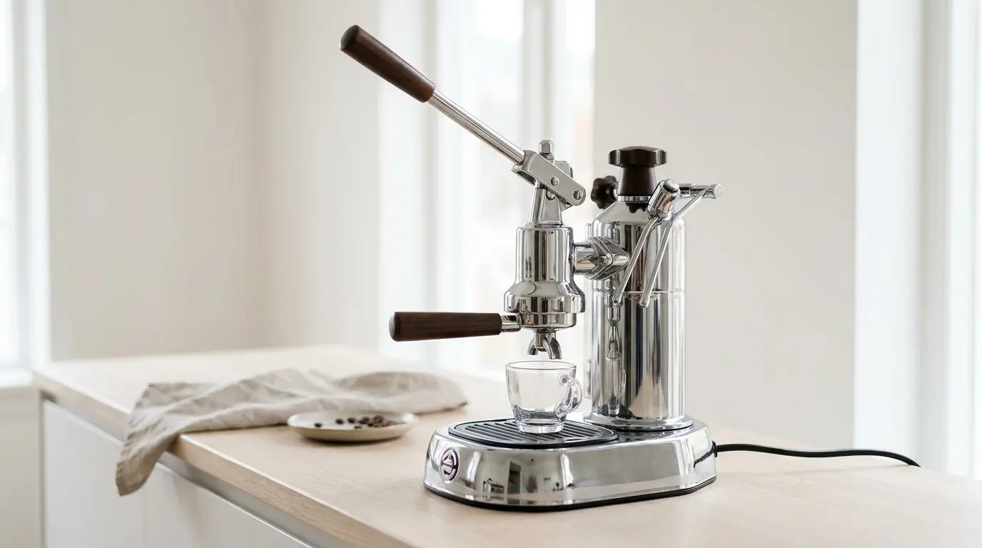 A polished chrome lever espresso machine with a tall body and a long wooden-handled lever raised, standing on a pale wooden counter in soft window light, a small glass cup beneath the group