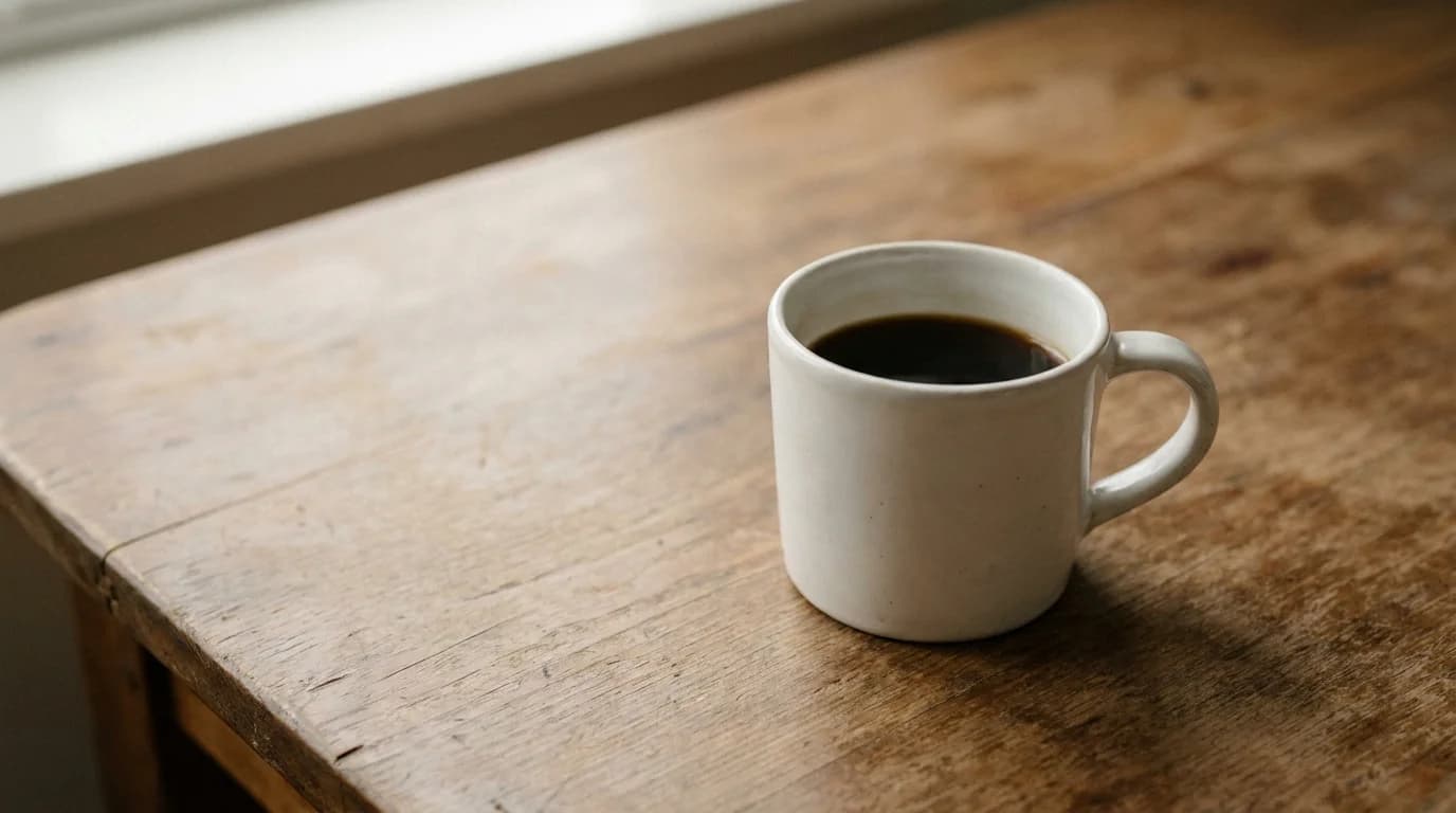A single white ceramic mug of dark coffee on a bare wooden surface with soft warm daylight, minimalist calm composition